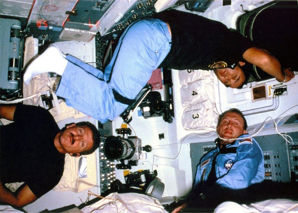 Group photo of three astronauts floating inside the space shuttle wearing blue pants and dark blue shirts.