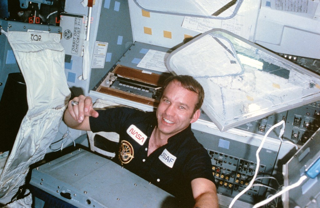 Image of an astronaut, visible from the chest up, wearing a black polo shirt, floating inside the space shuttle.