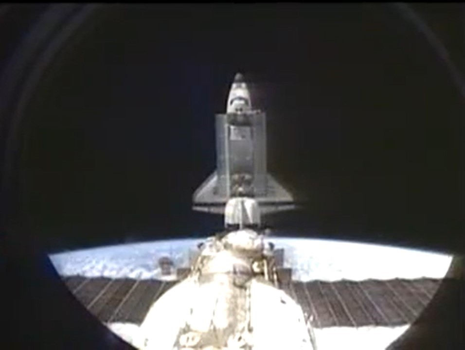 Image of a space shuttle against a black sky, with parts of another spacecraft (Mir) visible in foreground.