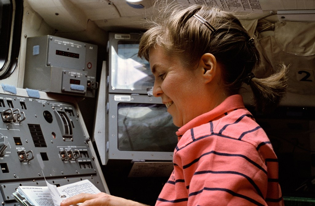 Image of a female astronaut wearing a pink and blue striped polo shirt operating equipment on the space shuttle's flight deck.