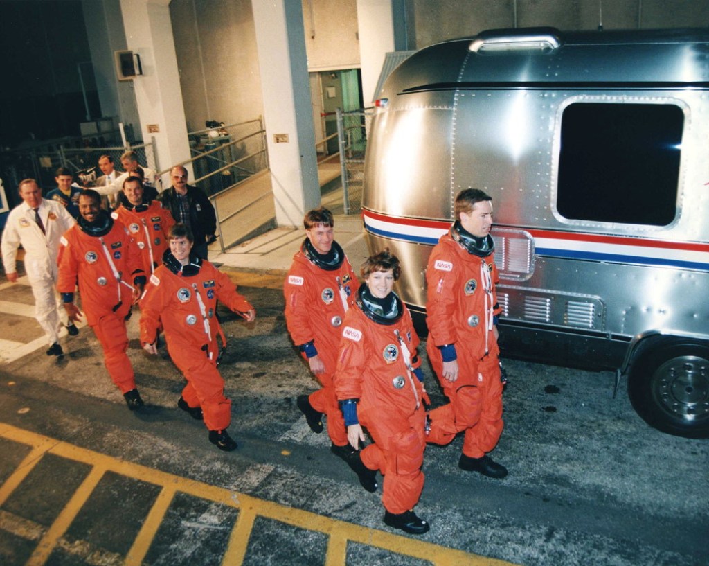 Group photo of six astronauts wearing orange spacesuits walking in front of a silver van.