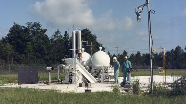 two men in slate gray coveralls stand at an early version of the test stand at the Diagnostic Testbed Facility