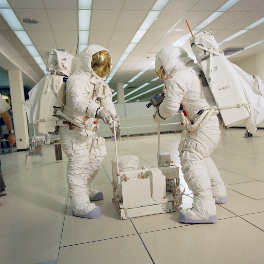 Two astronauts wearing white spacesuits practice setting up an experiment package during lunar surface training.