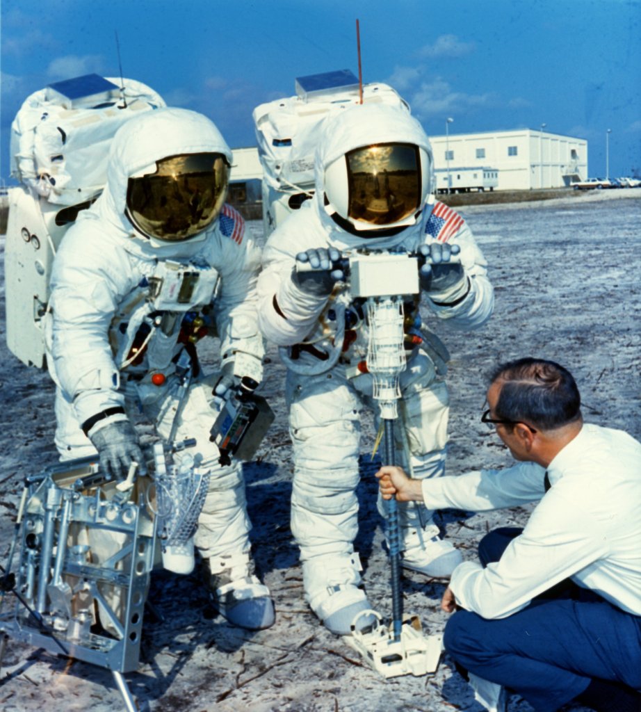 Two astronauts wearing white spacesuits practice using a drill during moonwalk training, assisted by a man in a white shirt.