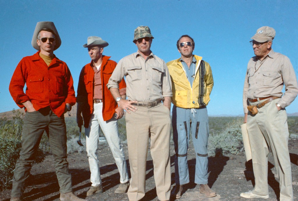 Image of five men, four astronauts and a geologist, standing in rugged terrain, against a blue sky background.