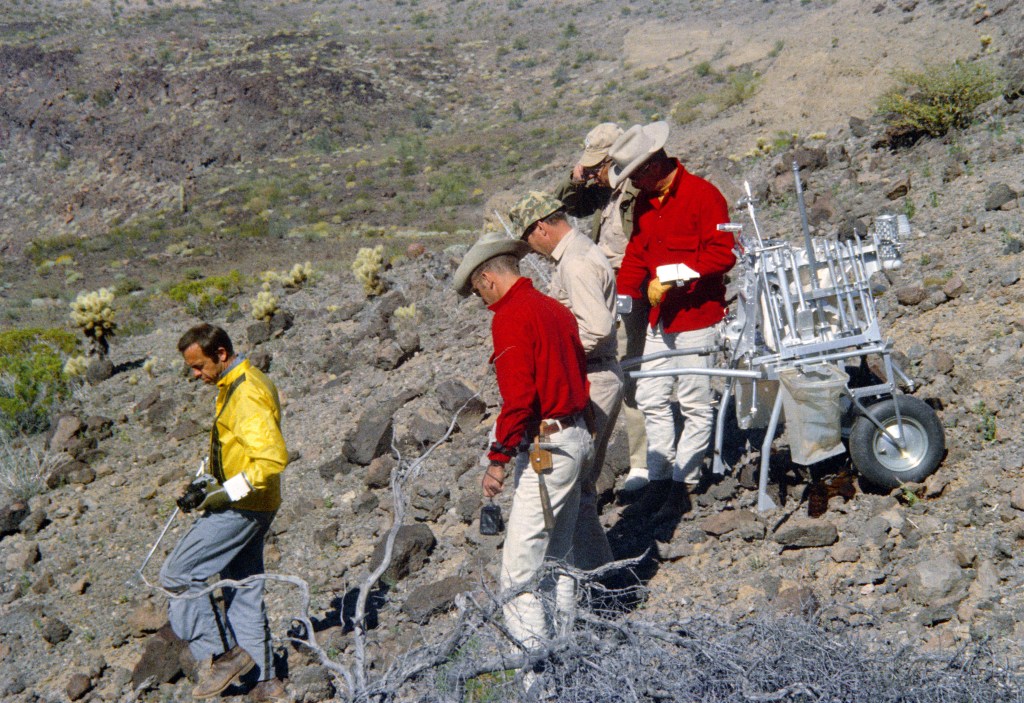 Image of five men, four astronauts and a geologist, working in rugged terrain, one of them pulling a golf cart like vehicle loaded with tools.