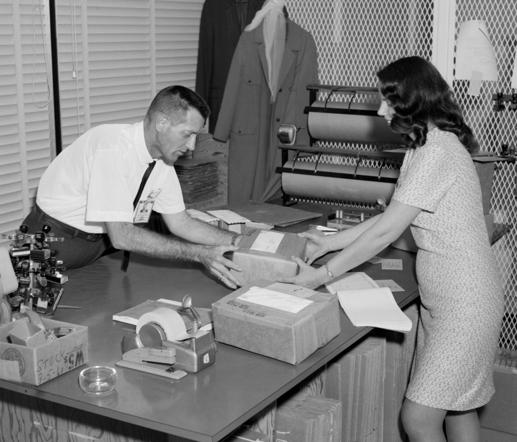 Black and white image of a man and a woman at a desk preparing packages for shipment.