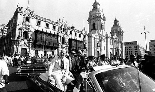 Black and white photo of three astronauts riding in a convertible with an ornate building in the background.