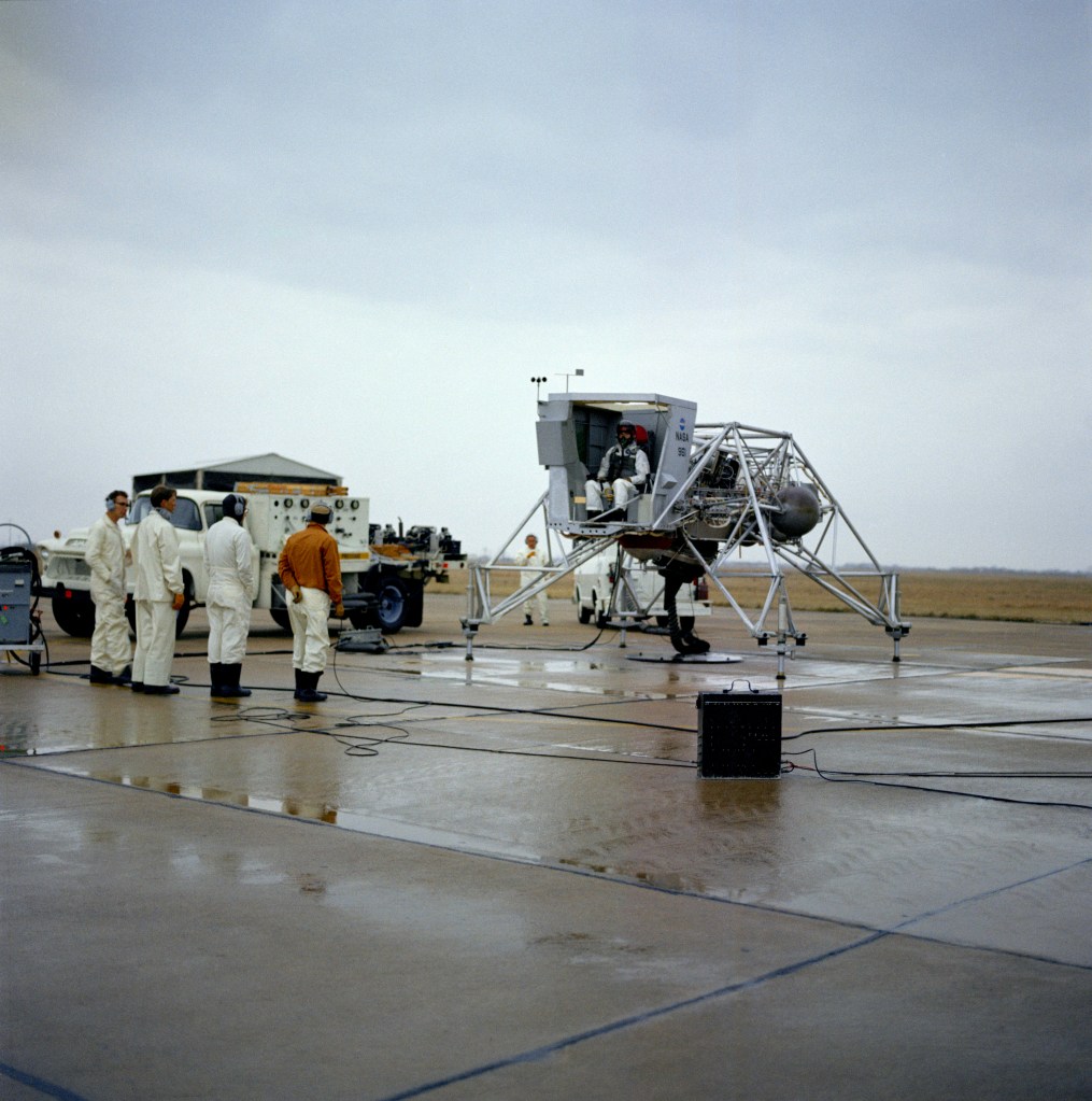 Scene on a runway of an astronaut wearing white overalls and a helmet sitting in the cockpit of the Lunar Landing Training Vehicle with several other men standing nearby in support.