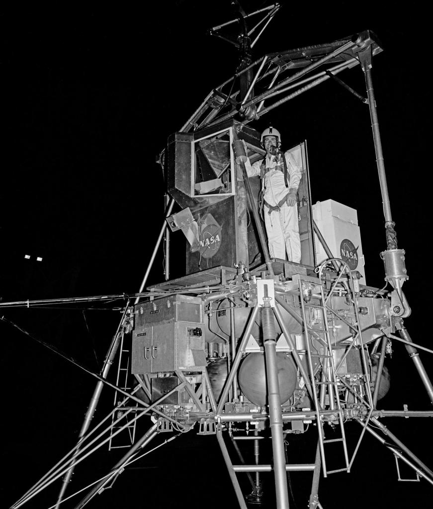 Nighttime scene of an astronaut in white overalls and a helmet standing in the cockpit of a lunar module trainer.