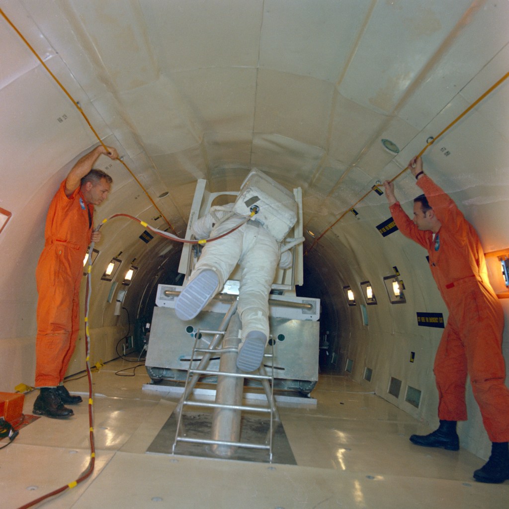 Image aboard an aircraft of white spacesuited astronaut practicing backing out of a lunar module mockup, with two men in orange overalls assisting him.