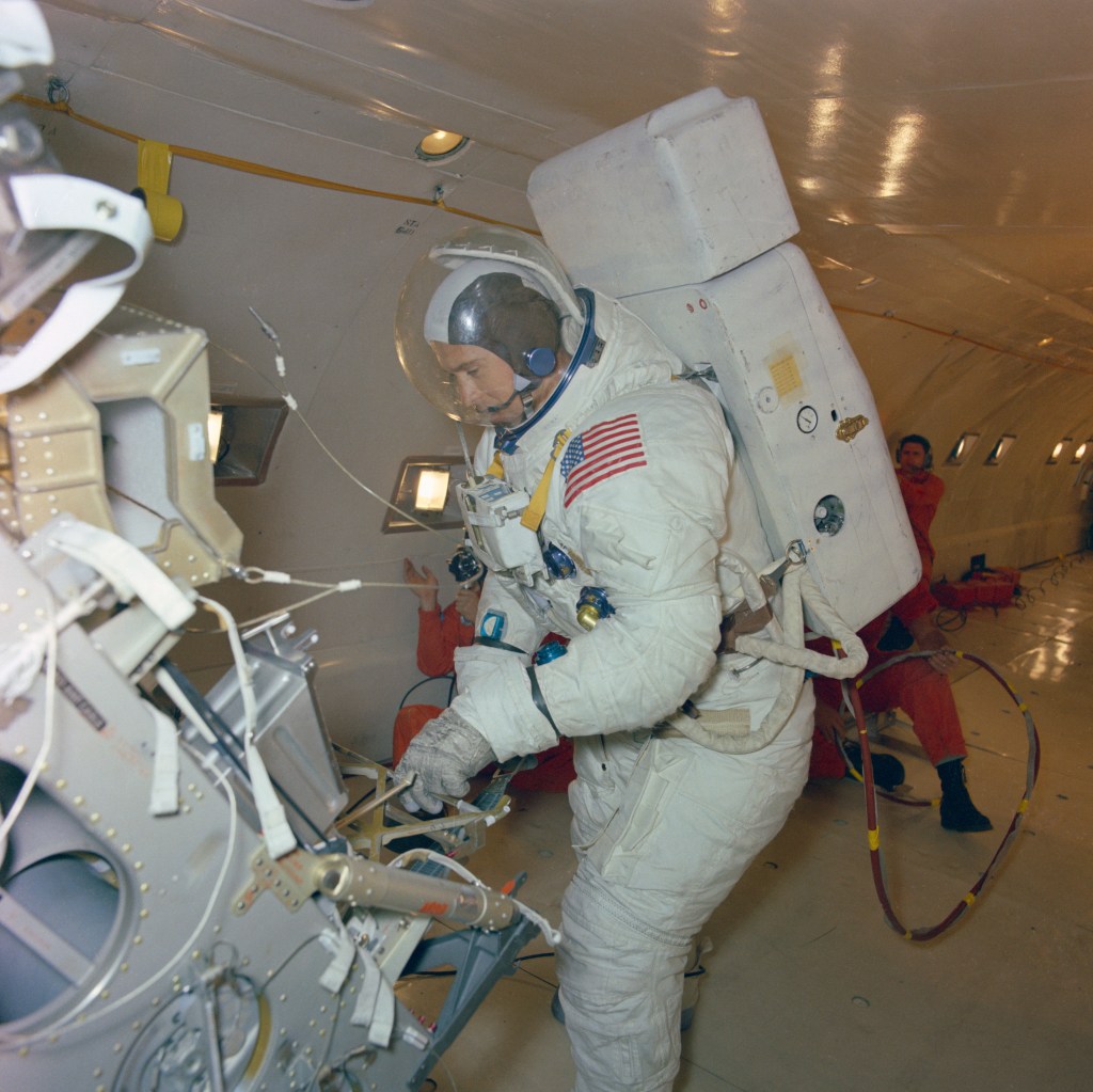 Image aboard an aircraft of a white spacesuited astronaut practicing unloading equipment from a lunar module mockup.