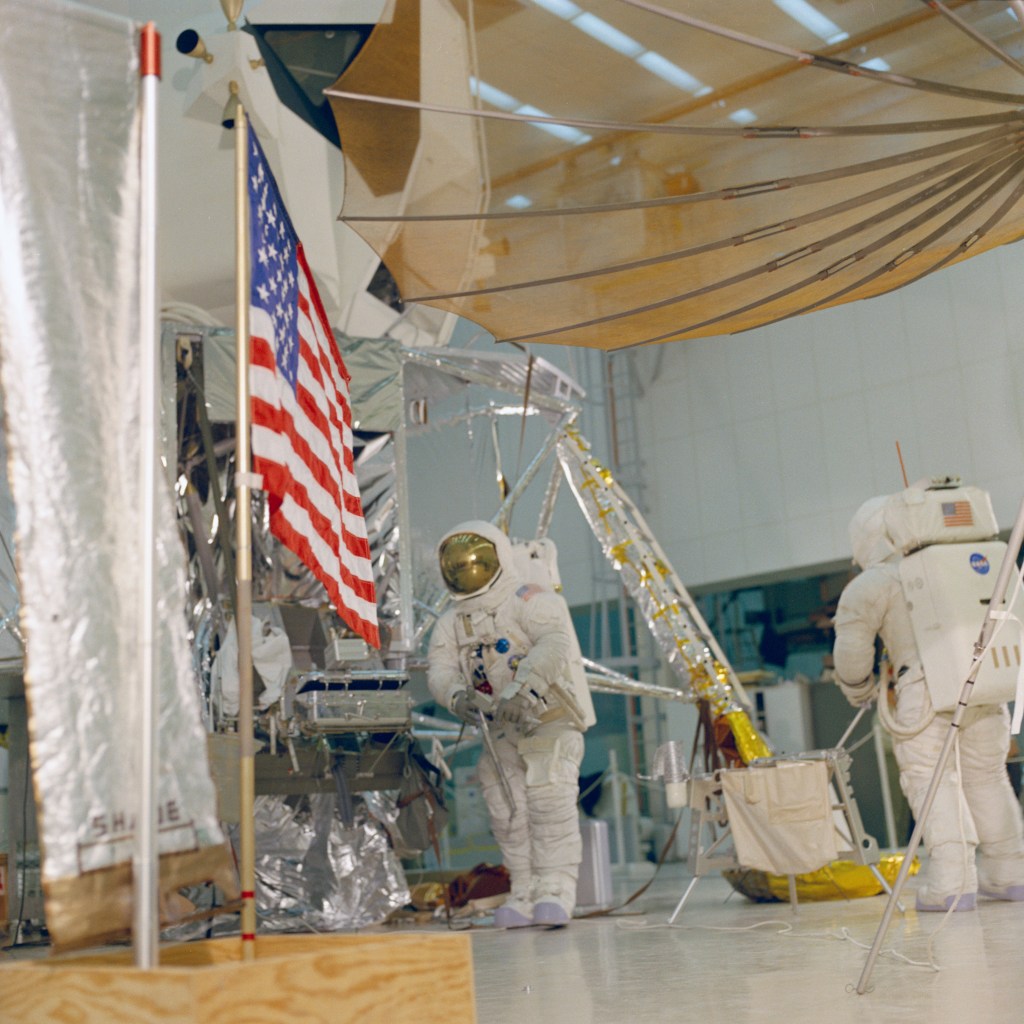 Two astronauts wearing white spacesuits practice a moonwalk, with an American flag visible in front of a lunar module mockup.