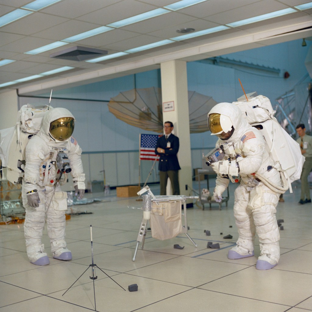 Two astronauts wearing white spacesuits practice a moonwalk and collecting rock samples.