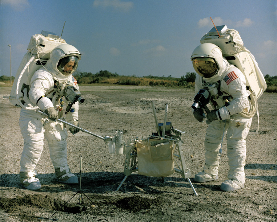 Two astronauts wearing white spacesuits practice setting up instruments in a moonwalk training exercise.