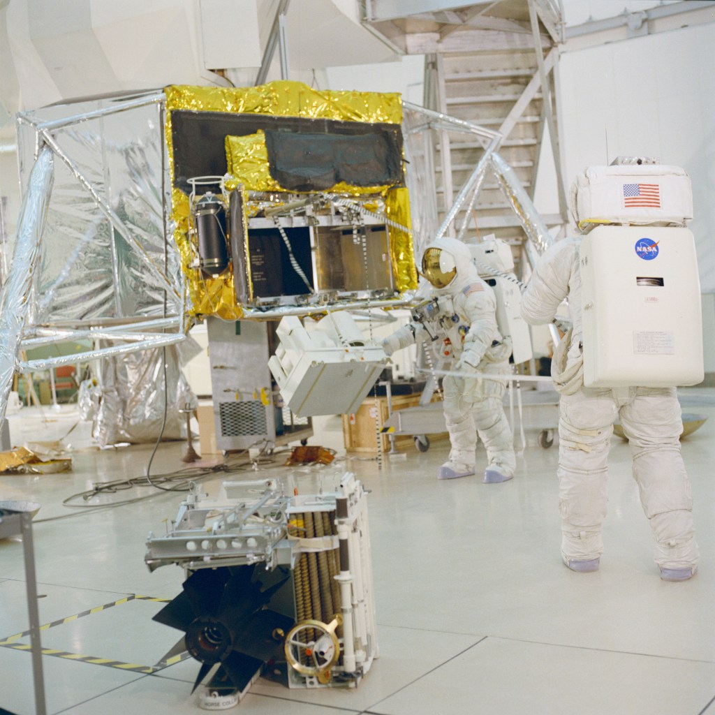 Two astronauts wearing white spacesuits practice removing equipment from a lunar module mockup during moonwalk training.