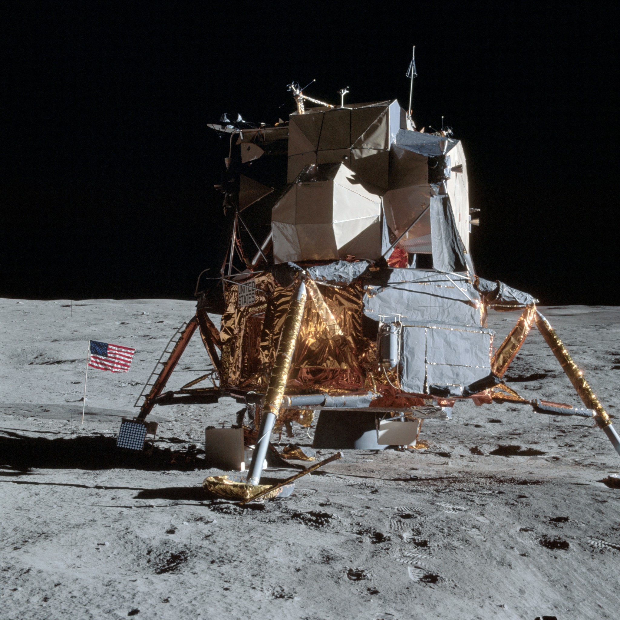 The Apollo 14 lunar module sits on the dusty and rocky gray surface of the Moon. The lunar module has a wide lower portion that is mostly covered in gold foil. Three of its legs are visible. The U.S. flag is next to the lunar module, at left. It is much shorter than the lunar module.