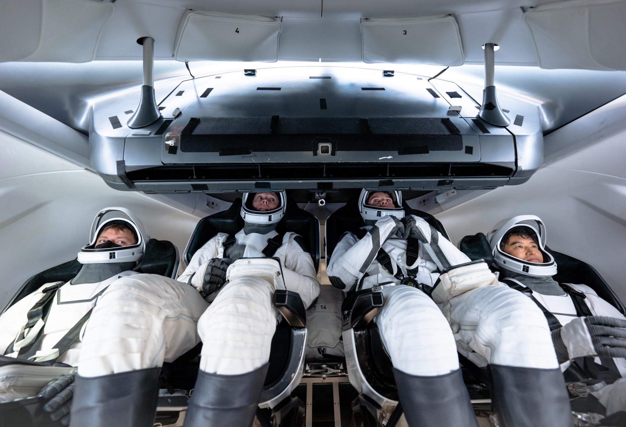 Four NASA SpaceX Crew-10 members sit in a capsule in their white spacesuits, looking around the spacecraft during training.