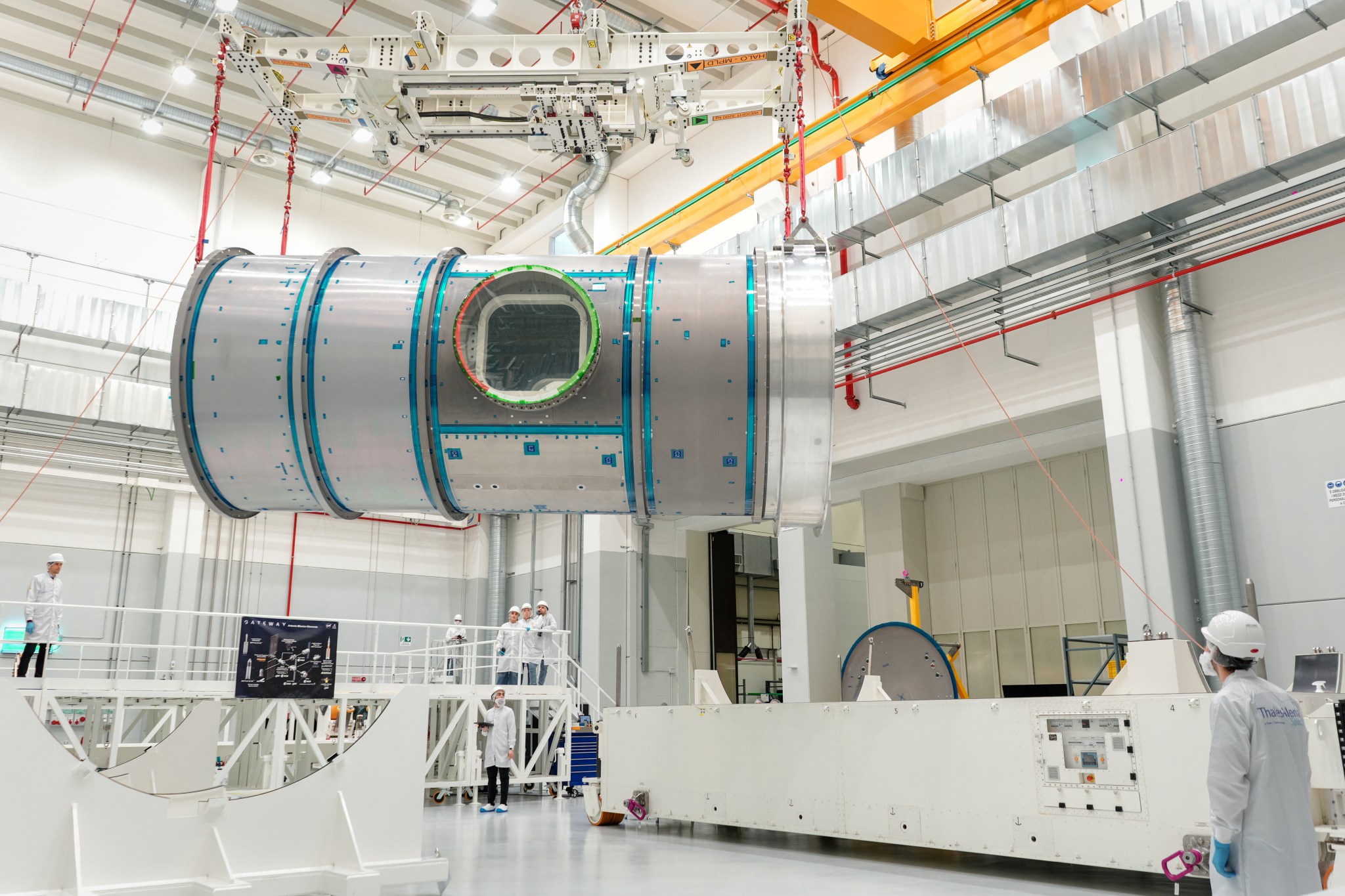 Gateway's Habitation and Logistics Outpost, a large cylindrical module, is suspended by red straps in a cleanroom at Thales Alenia Space in Turin, Italy. Engineers and technicians in white lab coats and helmets observe and guide the process. The cleanroom features metal walkways and bright overhead lighting.