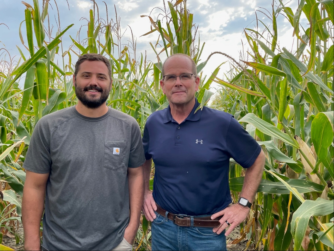 A picture of two white men standing in a cornfield. The one on the left is mid-thirties, with dark brown hair and beard and wearing a gray t-shirt. The man on the right is older, clean-shaven and wearing glasses and a navy blue collared shirt.