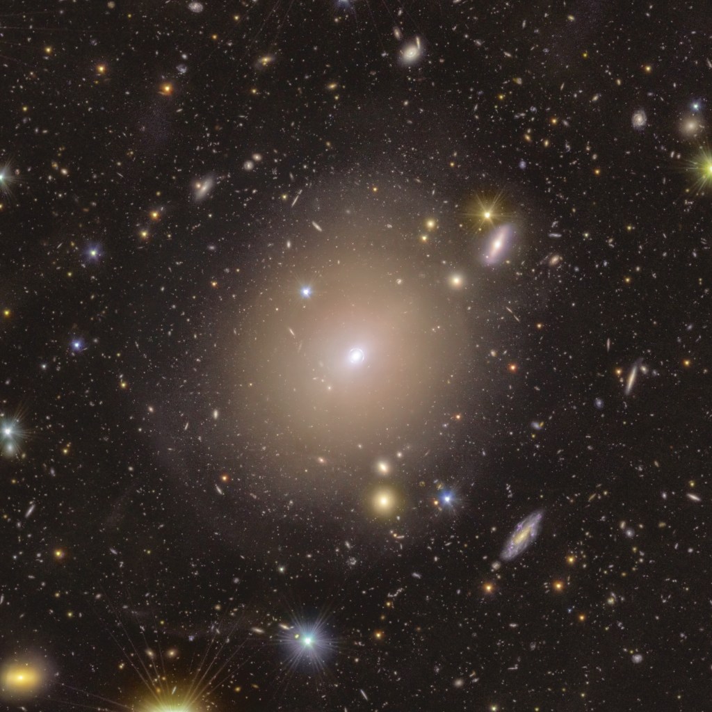 A sea of colorful stars and galaxies appear to swim in the vast blackness of space around a hazy halo at centre stage. In the middle of the image, the fuzzy-looking bulb of light in a warm shade of yellow extends around a small bright spot, nestled within a thin light circle that appears to be drawn closely around it. As we follow the central halo’s rim outwards, its brightness dims and blends smoothly into its surroundings. Here, extended discs of shades ranging from a warm purple to golden yellow, and piercing dots of light with sharp diffraction spikes are spread evenly across the image.