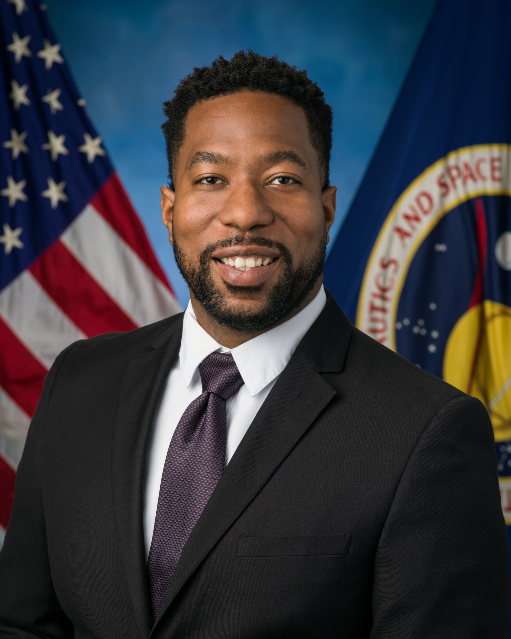 Professional headshot of a man wearing a dark suit and tie, smiling against a background featuring the United States flag and a NASA emblem.