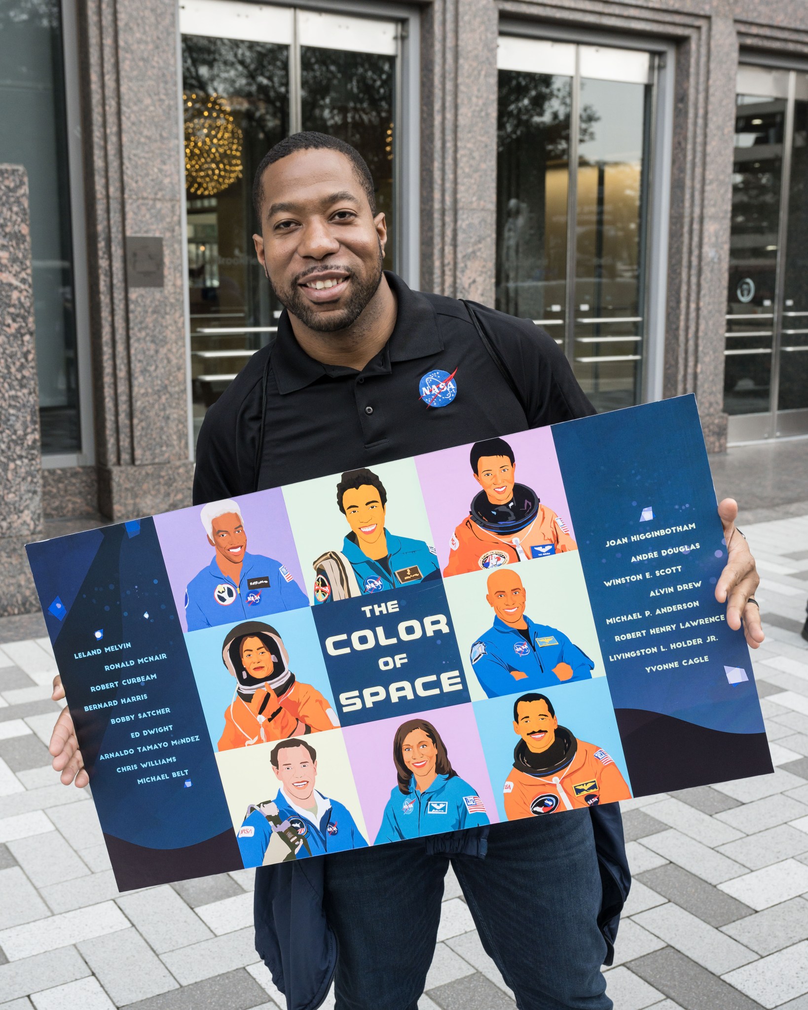 Man wearing a black NASA polo shirt, smiling and holding a colorful poster titled ’The Color of Space.’