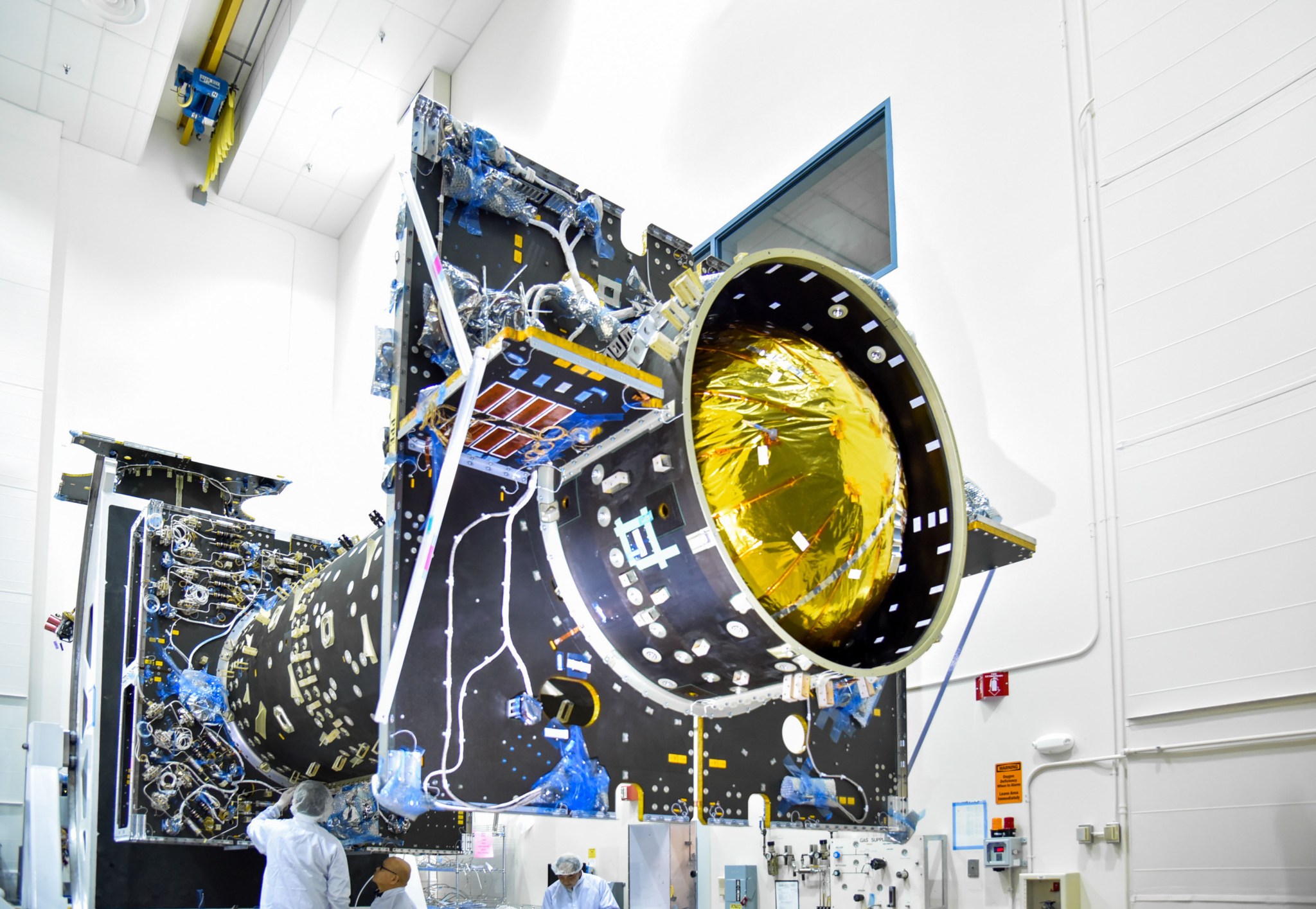 A side view of the Power and Propulsion Element for Gateway in a cleanroom at Maxar Space Systems in Palo Alto, California. The spacecraft is tilted at an angle, revealing a gold-colored tank housed inside the large cylindrical structure. The exterior is lined with wiring and connectors.  Technicians in cleanroom attire work below. 