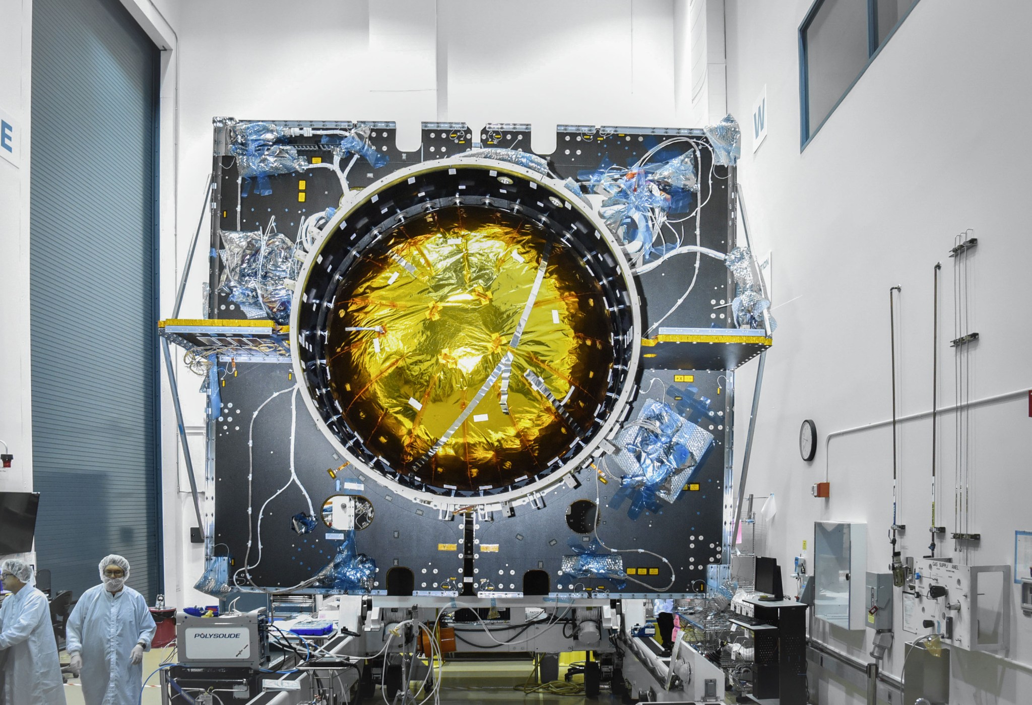 The Power and Propulsion Element of Gateway is shown being assembled inside a clean room at Maxar Space Systems in Palo Alto, California. The large cylindrical structure has numerous wires and components attached to its black exterior paneling. A reflective gold-colored fuel tank is seen inside the main clylinder. Engineers in cleanroom suits work around the spacecraft, inspecting and assembling its systems.