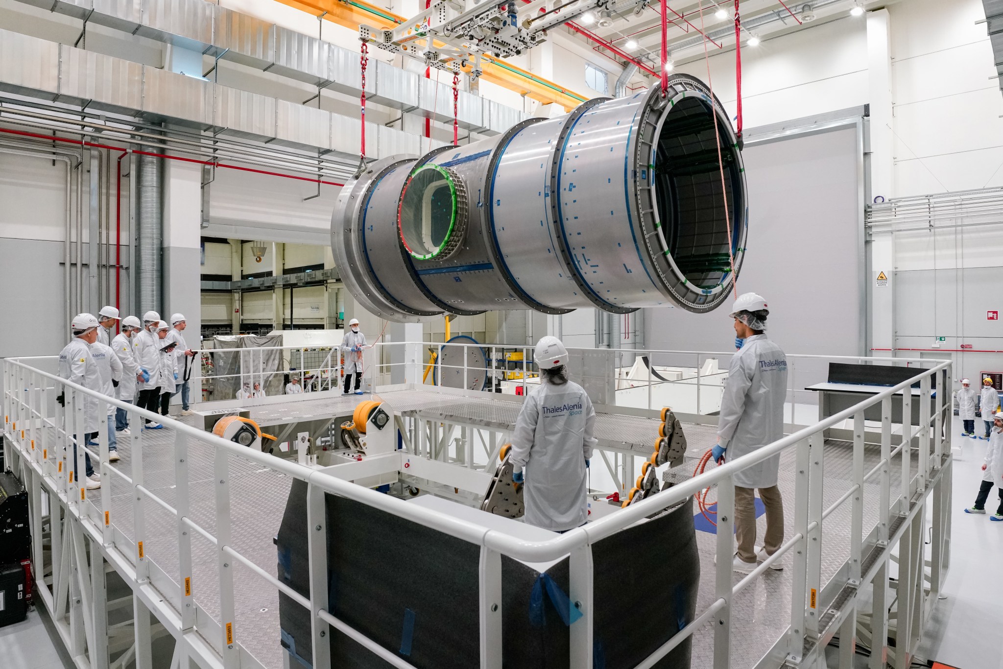 A large cylindrical module is suspended by red straps as it is lowered onto a stand in a cleanroom at Thales Alenia Space in Turin, Italy. Engineers and technicians in white lab coats and helmets observe and guide the process from an elevated platform. The cleanroom features metal walkways and bright overhead lighting.