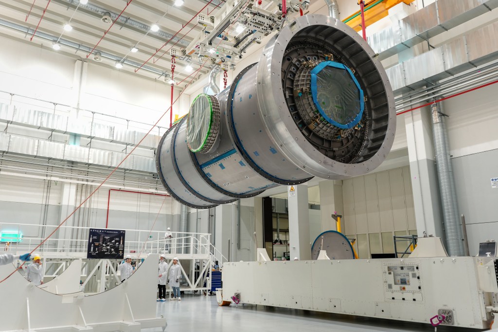 A large cylindrical module, Gateway's Habitation and Logistics Outpost, is suspended by red straps as it is lowered onto a stand in a cleanroom at Thales Alenia Space in Turin, Italy. Engineers and technicians in white lab coats and helmets observe and guide the process. The cleanroom features metal walkways and bright overhead lighting.