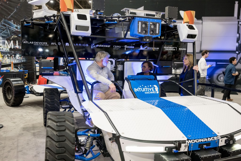 Three individuals interacting around a lunar rover by Intuitive Machines at an exhibit.