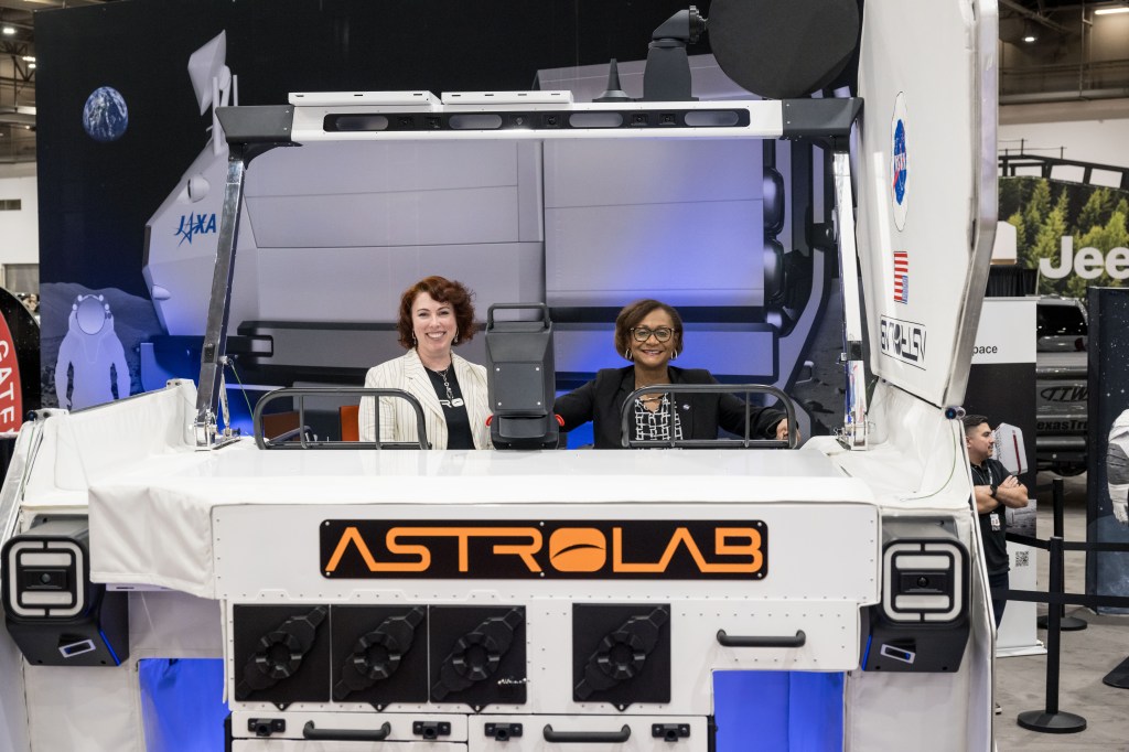 Two individuals seated inside an Astrolab rover at an exhibit, smiling for the camera.