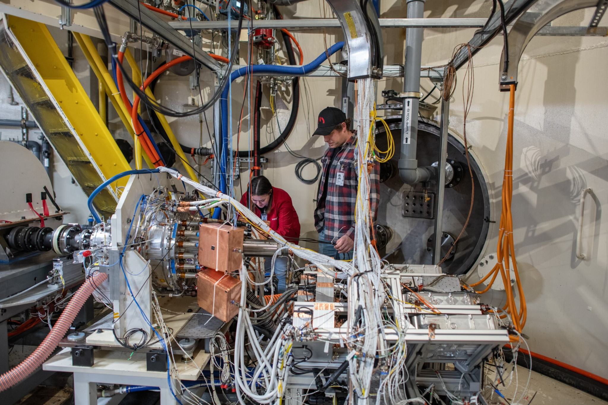 A man and a women set up an electric engine inside a large testing facility surrounded by various wires, cables, and equipment that will help simulate the effects of flight at high altitudes. 