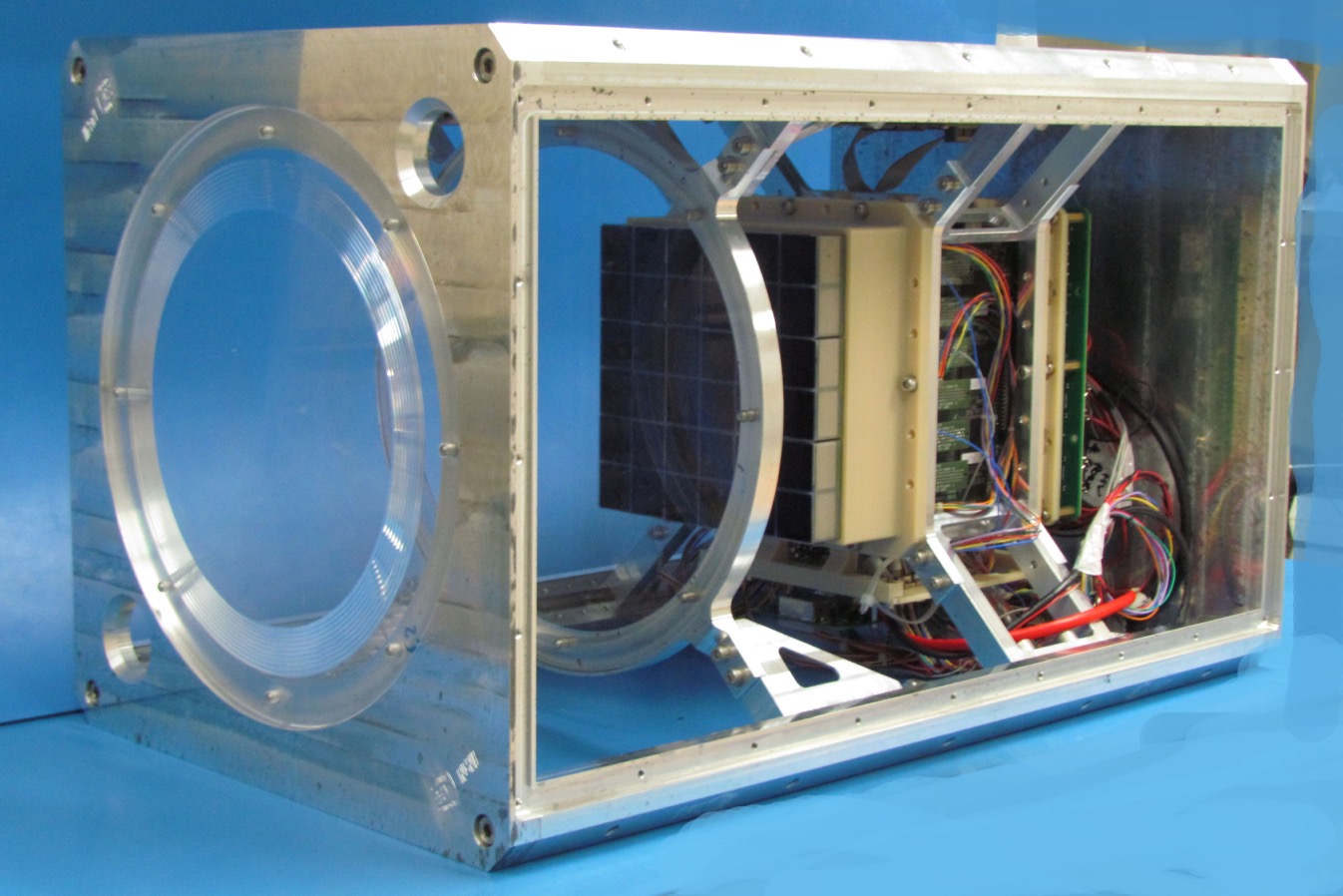 The Mini-EUSO rectangular casing is shown during assembly and the engineering hardware can be seen on the inside. Near the middle of the rectangular structure is a six-by-six grid—this is the photomultiplier, or light detecting mechanism of the telescope. Wires and brackets are connected to the back of the photomultiplier unit.