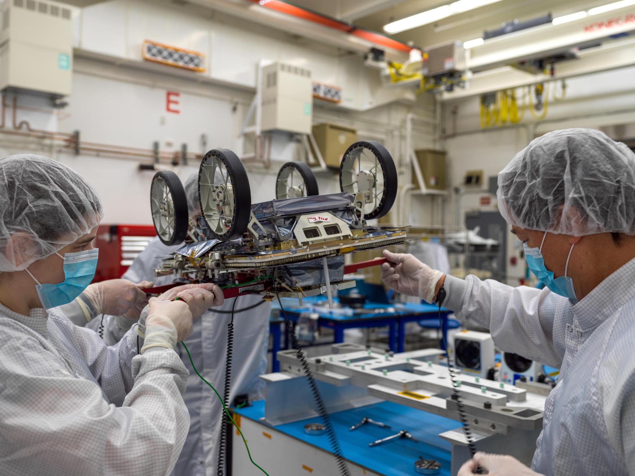 Four people in white lab coats, face masks, and hair nets hold up a small, upside-down robotic rover by red handles inside a room with industrial equipment in the background.