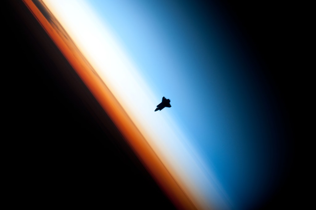 Image of a small space shuttle silhouetted against a multicolored limb view of the Earth's atmosphere.