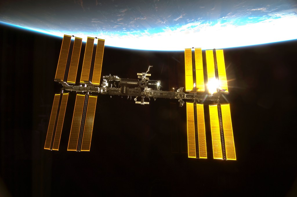 View of the International Space Station as seen from an approaching space shuttle, seen against the blackness of space and a thin crescent of Earth.