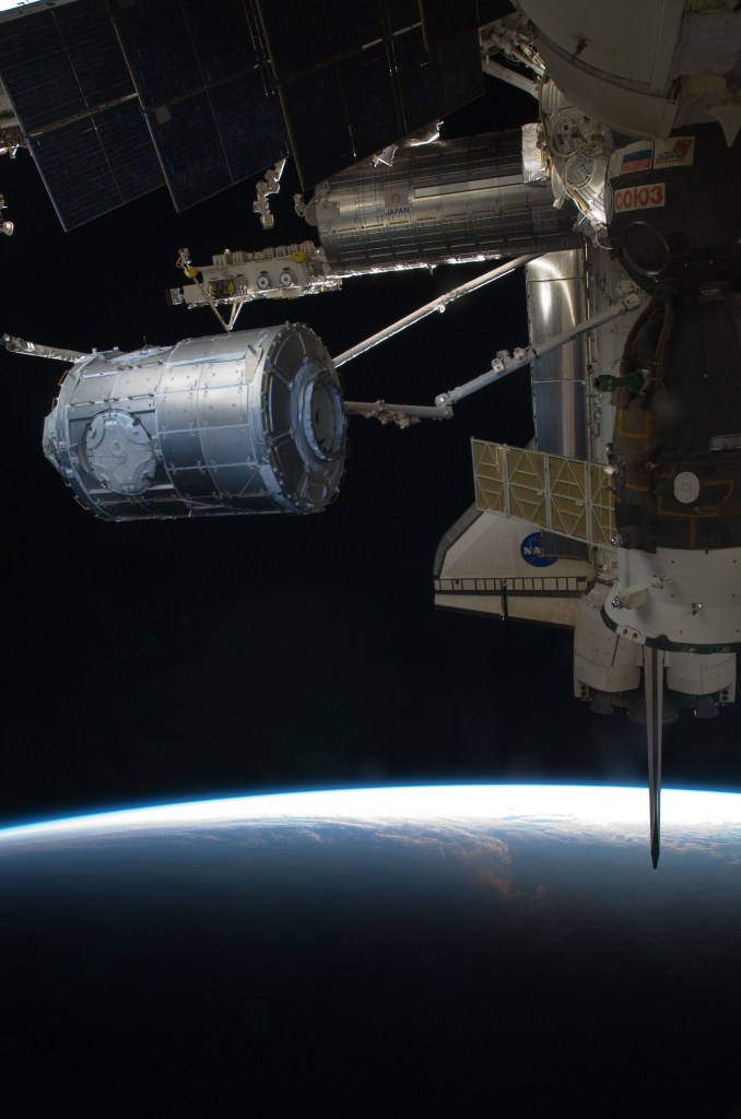 Image of the space station robotic arm moving a large cylindrical module from the space shuttle to the space station.