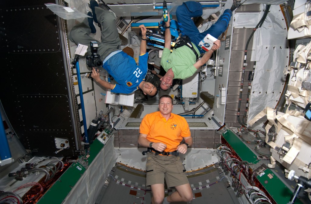 Three astronauts wearing orange, blue and green T-shirts float head-to-head inside the International Space Station.