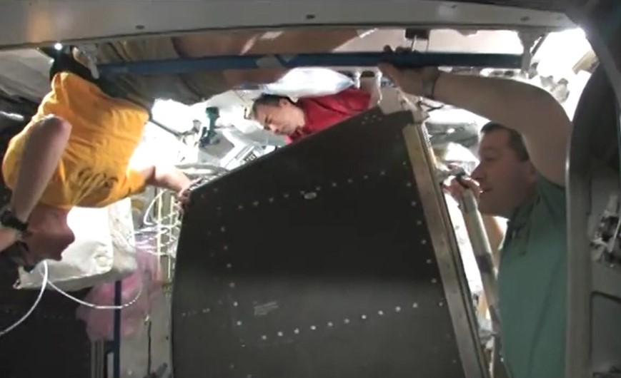 View of three male astronauts inside the space station moving a large rack.