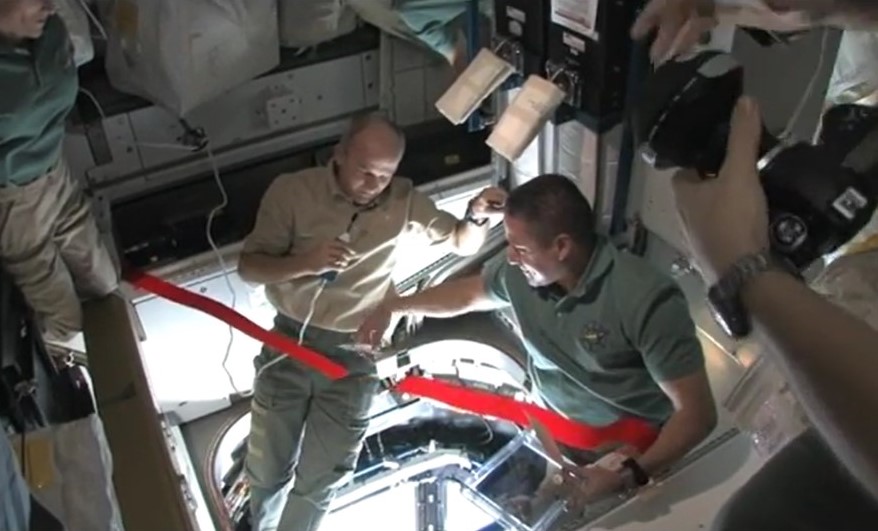 View of astronauts inside the space station cutting a red ribbon.