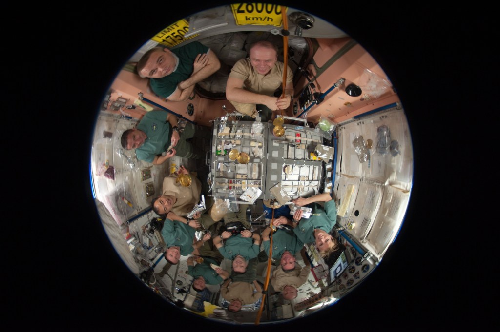 Fisheye lens view of 11 astronauts sharing a meal together inside the space station.