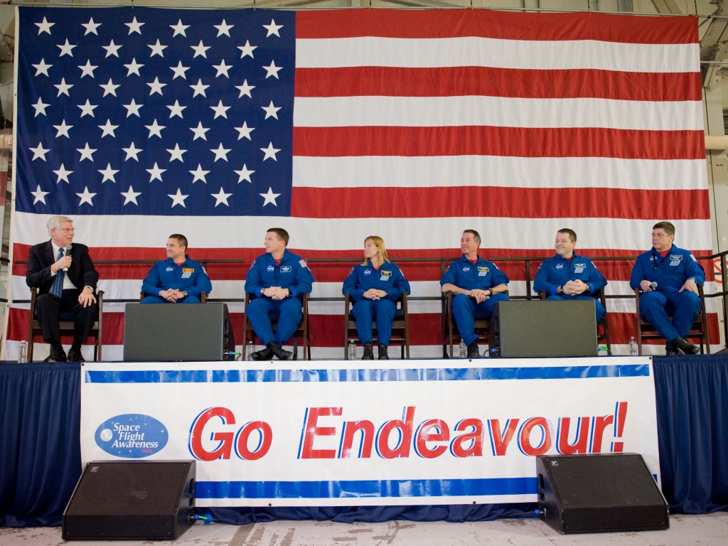 Six astronauts wearing blue overalls seated on a stage with a large American flag behind them.