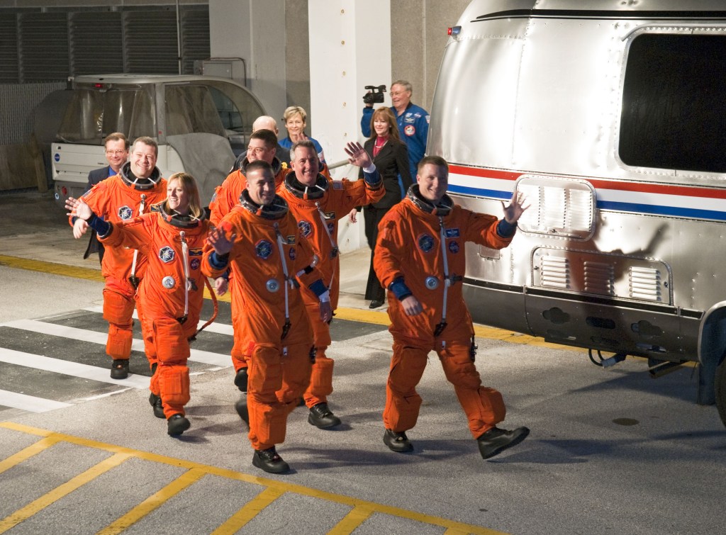 Six astronauts wearing orange spacesuits walkout of a building next to a van.