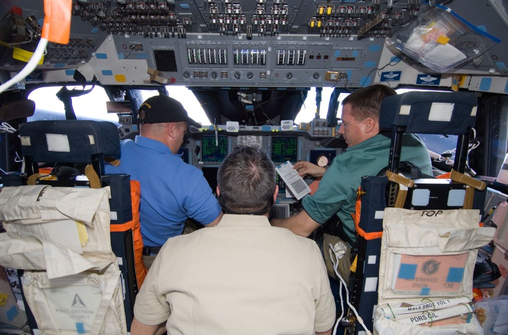 Image of three astronauts, backs to the camera, on the flight deck of the space shuttle.