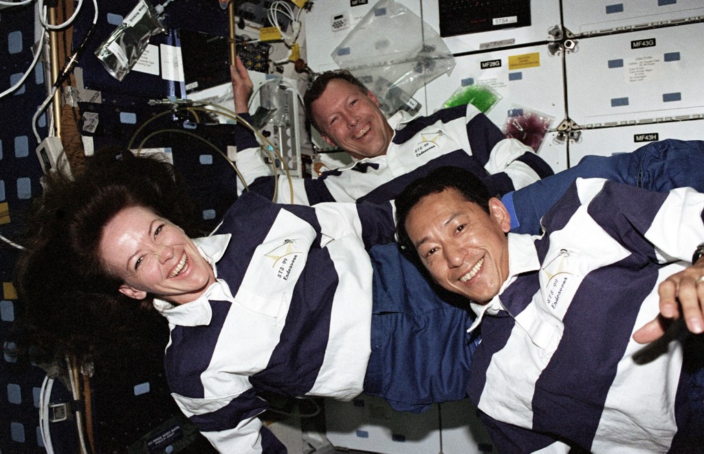 Image of one female and two male astronauts wearing white and blue striped jerseys floating in the shuttle middeck.