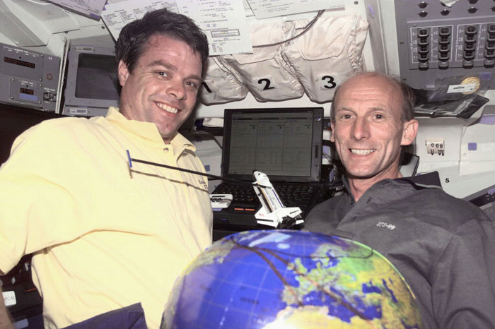 Image of two male astronauts, one wearing a yellow shirt, the other a gray one, with an inflatable globe of the Earth between them, floating on the shuttle's flight deck.