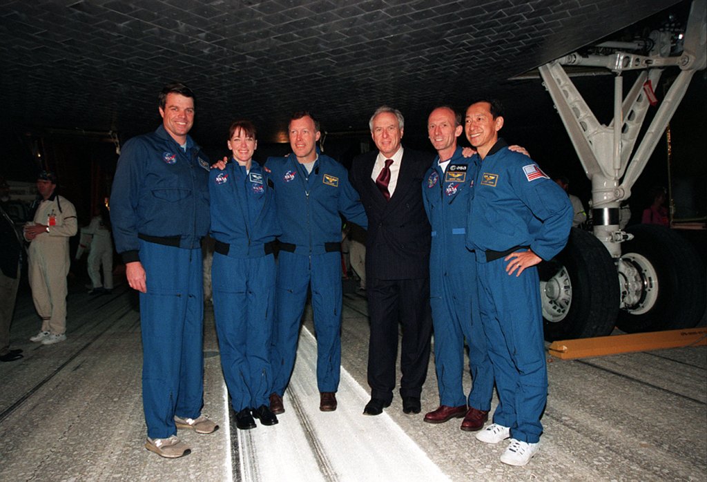 Five astronauts wearing blue overalls and one man in a black suit pose in front of a space shuttle.