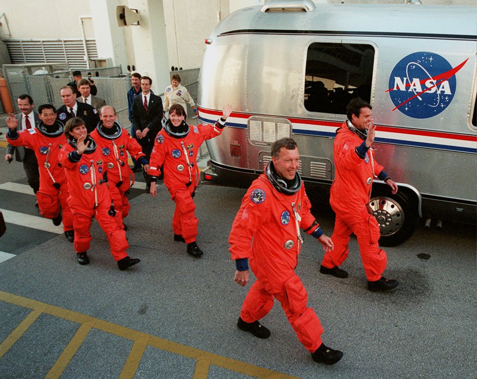 Image of six astronauts dressed in orange spacesuits walking in front of a silver van.